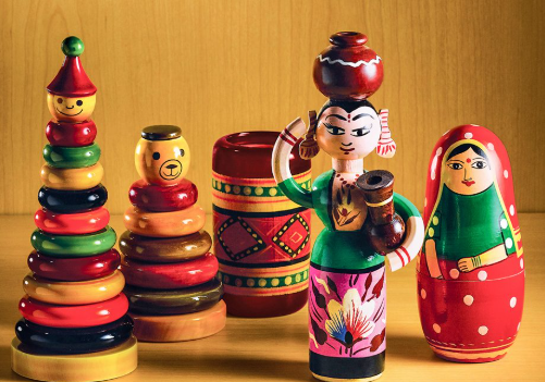 A collection of colorful wooden toys and figurines, including a stacked toy, a bear, a traditional woman holding a pot, and two nesting dolls.