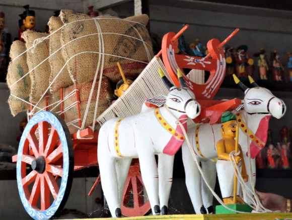 A colorful traditional wooden cart pulled by two white bulls, filled with burlap sacks. A small figure representing a farmer stands beside the cart, with various handmade toys in the background.