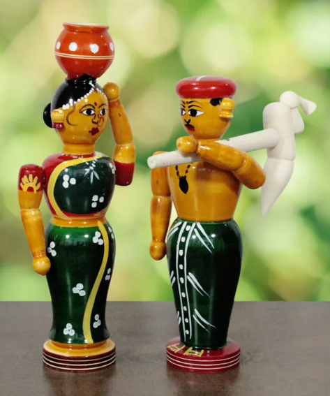 Two colorful wooden figurines representing a traditional couple, one woman with a pot on her head and one man holding a farming tool, set against a blurred green background.
