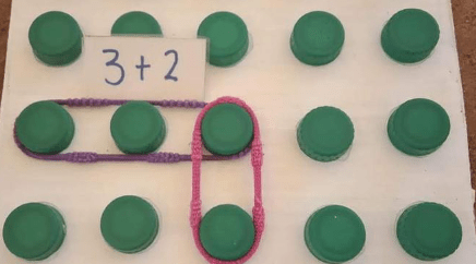 A math activity using green bottle caps arranged on a white board, with a card displaying '3+2' and rubber bands connecting certain caps to illustrate addition.
