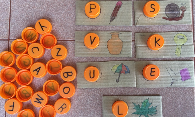 A collection of orange bottle caps with letters arranged in a circular pattern next to illustrated cardboard cards featuring drawings of a pencil, snail, vase, key, umbrella, and leaf, each topped with a matching orange cap.
